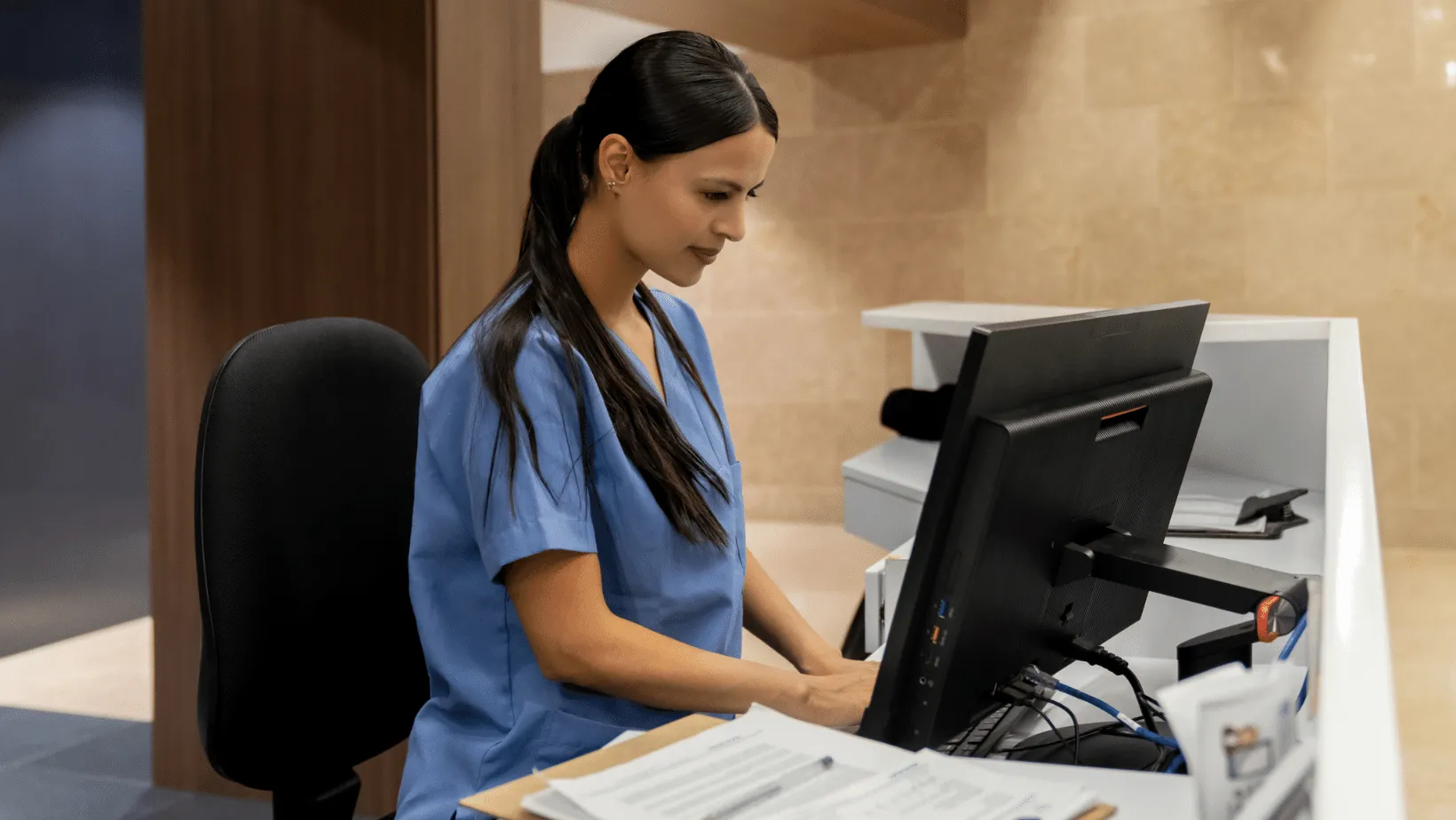 A focused healthcare worker in blue scrubs working at a computer station, presumably in a medical office or hospital setting, with papers and a phone visible on the desk.