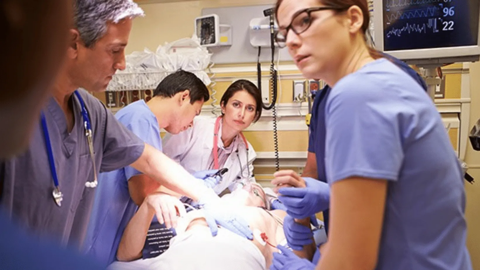 A team of medical professionals in scrubs and protective gear intently performing a procedure on a patient in an emergency room, with medical monitors in the background.