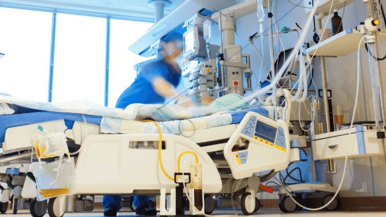 Motion blur image of a nurse in blue scrubs attending to a patient in an intensive care unit (ICU), surrounded by advanced medical equipment and monitors.