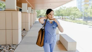 A healthcare worker in blue scrubs is putting on a face mask outside a hospital, with a brown shoulder bag and a badge around her neck, walking along a covered pathway.